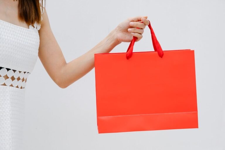 A fashionable woman in a white dress holds a bold red shopping bag against a neutral background.
